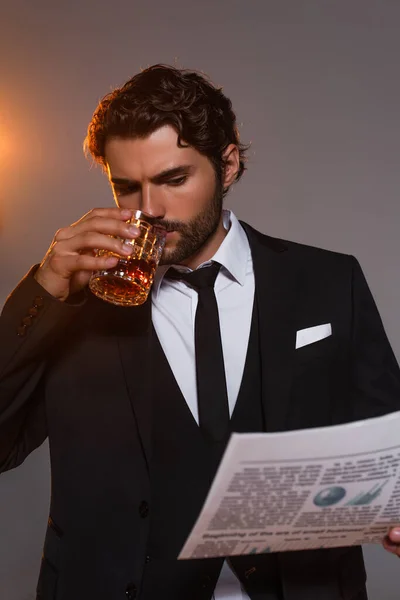 Hombre serio en chaqueta negra leyendo periódico y bebiendo whisky sobre fondo gris con luz - foto de stock