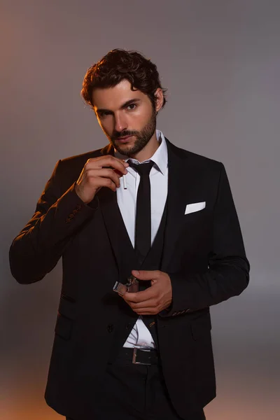 Bärtiger Mann im schwarzen Anzug blickt in die Kamera, während er Zigarette und Feuerzeug auf grauem Hintergrund hält — Stockfoto