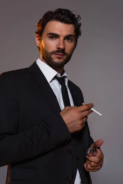 Стильный мужчина в черном костюме смотрит в камеру, держа в руках сигарету и зажигалку, изолированную на сером — стоковое фото