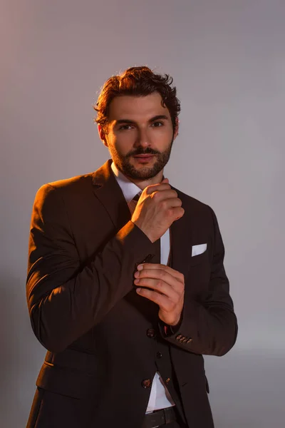 Elegant man buttoning sleeve of black blazer while looking at camera isolated on grey — Stock Photo