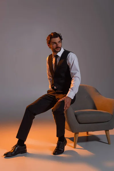 Vista completa del elegante hombre de negocios mirando hacia otro lado cerca del sillón sobre fondo gris con luz - foto de stock