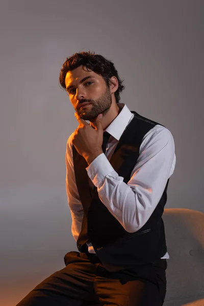 Stylish brunette man in white shirt and black vest touching chin and looking at camera on grey background — Stock Photo