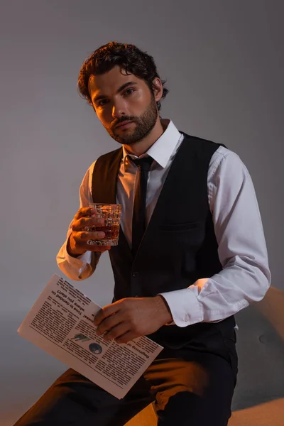 Homme élégant avec verre de whisky et journal regardant la caméra sur fond gris — Photo de stock