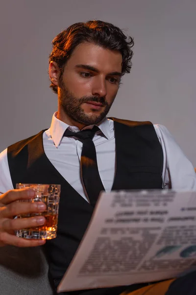 Brünetter Geschäftsmann hält ein Glas Whiskey in der Hand und liest Zeitung isoliert auf grau — Stockfoto