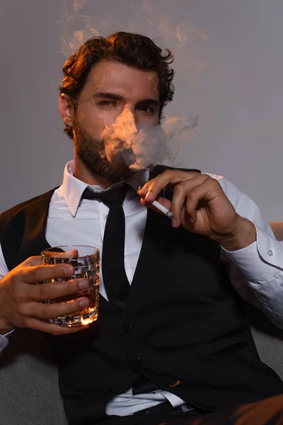 Homme élégant avec un verre de whisky cigarette fumante et regardant la caméra sur fond gris — Photo de stock