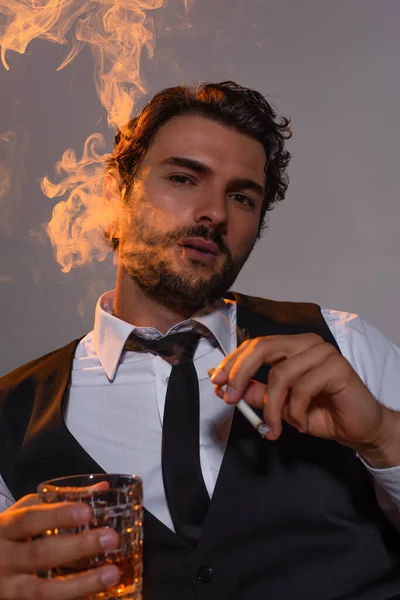 Homme élégant avec verre de whisky regardant l'appareil photo et fumant sur fond gris avec de la lumière — Photo de stock