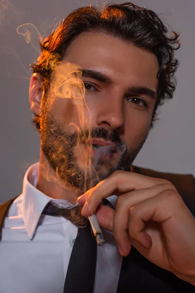 Vista de cerca del hombre con estilo que mira una cámara a través del humo del cigarrillo sobre fondo gris - foto de stock