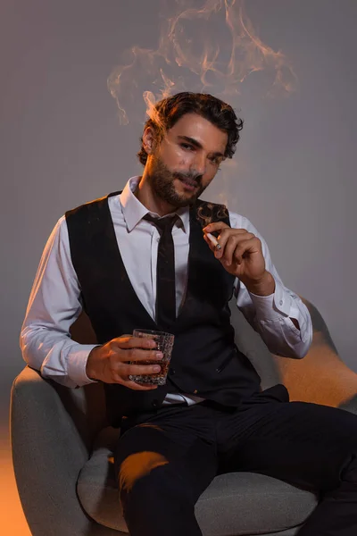 Элегантный брюнетка мужчина со стаканом виски сидит в кресле и курит на сером фоне — стоковое фото