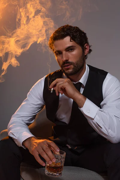 Елегантний чоловік сидить у кріслі зі склянкою віскі та цигарки на сірому фоні з димом та світлом — стокове фото