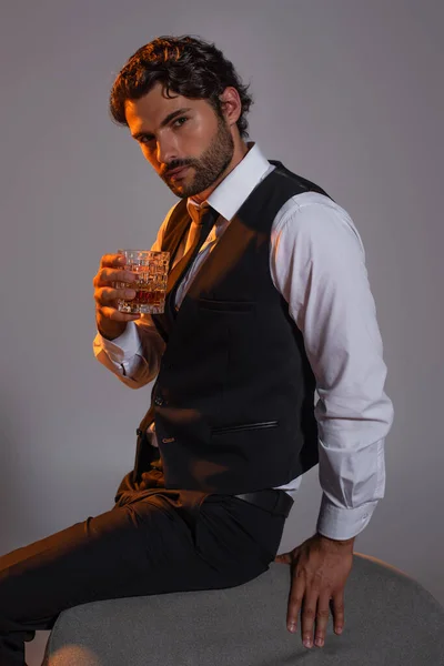 Uomo in gilet nero e camicia bianca guardando la fotocamera mentre tiene il bicchiere di whisky isolato sul grigio — Foto stock