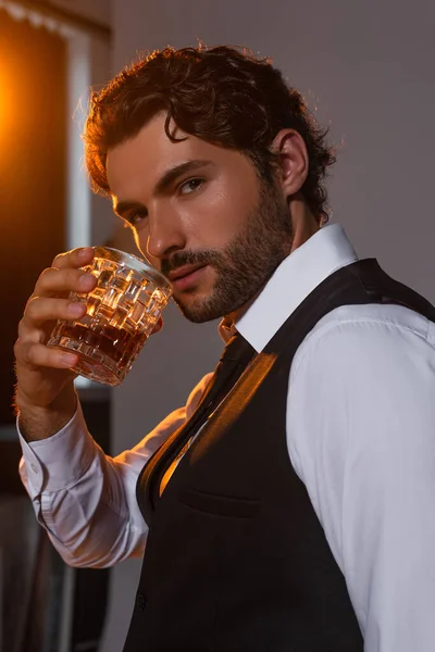 Brunette man in white shirt and black vest holding glass of whiskey on grey background with light — Stock Photo