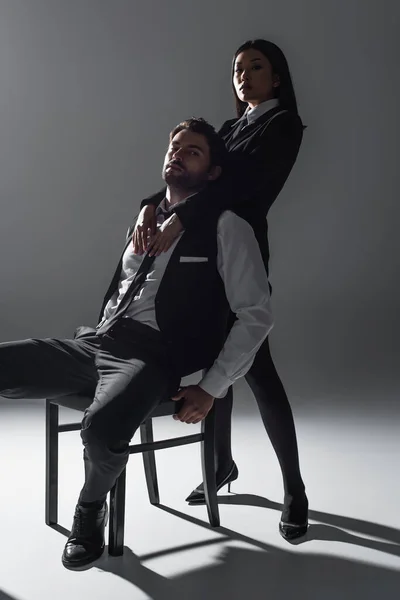 Stylish man sitting on chair near sexy asian woman seducing him on grey background — Stock Photo