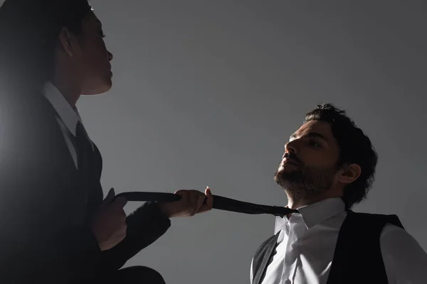 Leidenschaftliche asiatische Frau mit Krawatte eines brünetten Mannes, während sie ihn isoliert auf grau verführt — Stockfoto