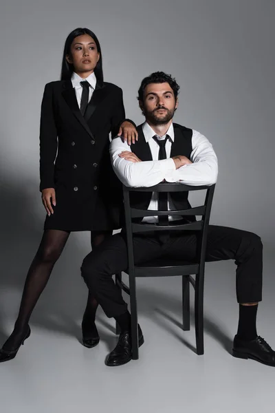 Sexy asiatische Frau im schwarzen Blazerkleid in der Nähe eleganter Mann sitzt auf Stuhl auf grauem Hintergrund — Stockfoto