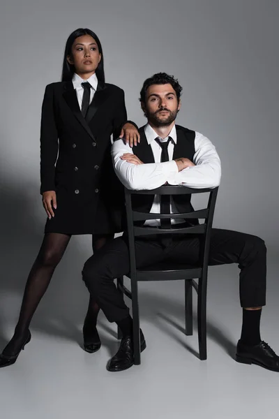 Sexy asiático mujer en negro blazer vestido cerca elegante hombre sentado en silla en gris fondo - foto de stock