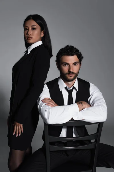 Elegant man sitting on chair with crossed arms near sexy asian woman on grey background — Stock Photo