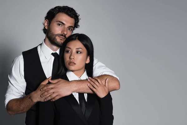 Elegante hombre abrazando morena asiático mujer y mirando cámara en gris fondo - foto de stock