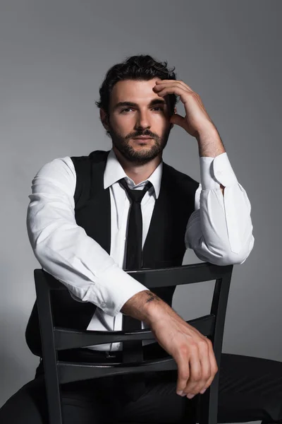 Stylischer Mann in schwarzer Weste und Krawatte sitzt auf Stuhl und blickt vereinzelt in die Kamera auf grau — Stockfoto