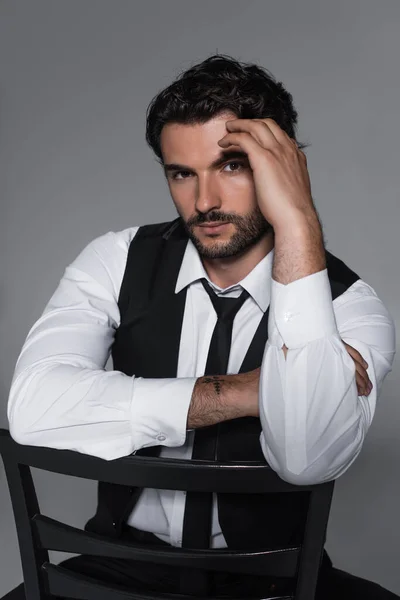 Stylish man in black vest and tie looking at camera while sitting isolated on grey — Stock Photo