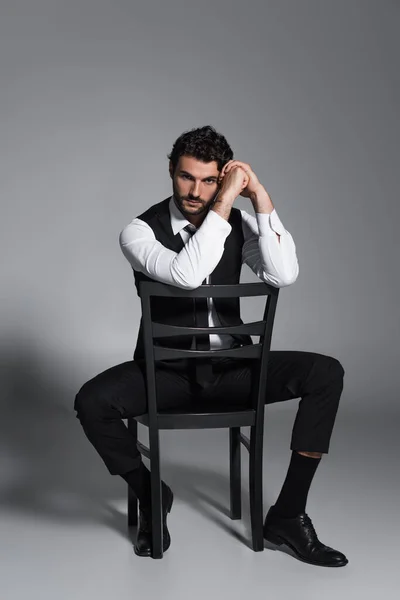 Full length view of brunette man sitting on chair and looking at camera on grey — Stock Photo