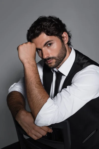 Brunette man in black vest and white shirt looking at camera while sitting isolated on grey — Stock Photo