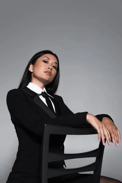 Sensual asian woman in black blazer and tie posing on chair isolated on grey — Stock Photo