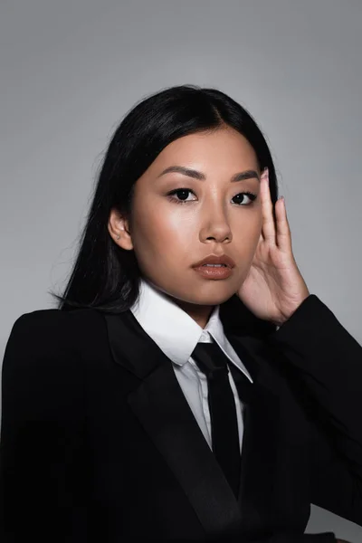 Morena mujer asiática en negro chaqueta y corbata mirando cámara aislada en gris - foto de stock