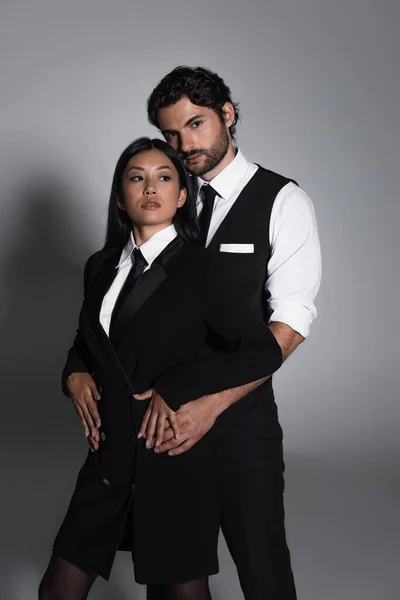 Brunette man looking at camera while embracing asian woman in black blazer dress on grey background — Stock Photo