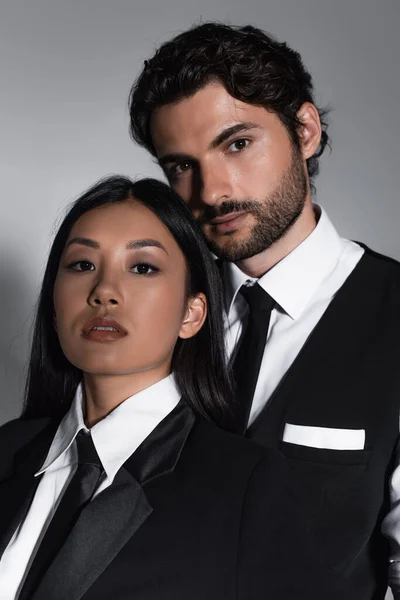 Bruna donna asiatica ed elegante uomo barbuto guardando la fotocamera su sfondo grigio — Foto stock