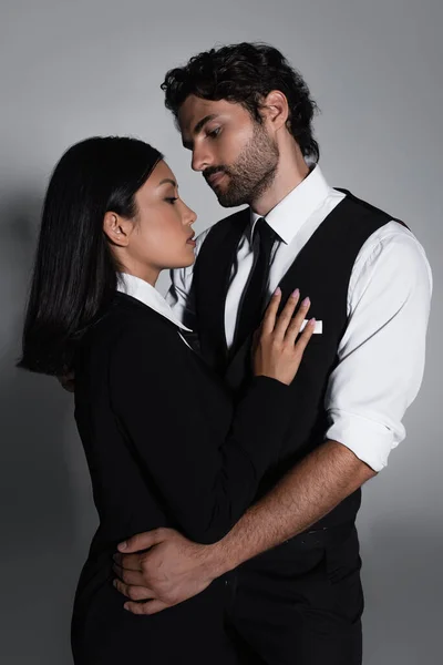 Morena hombre en formal desgaste abrazando bastante asiático mujer en gris fondo - foto de stock