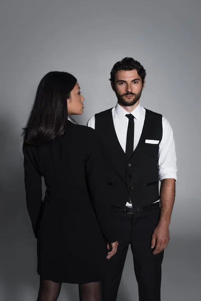 Vista trasera de la mujer asiática en vestido de chaqueta negro cerca de hombre elegante mirando a la cámara sobre fondo gris - foto de stock