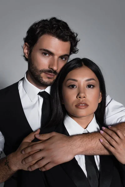 Brunette man embracing sensual asian woman while looking at camera isolated on grey — Stock Photo