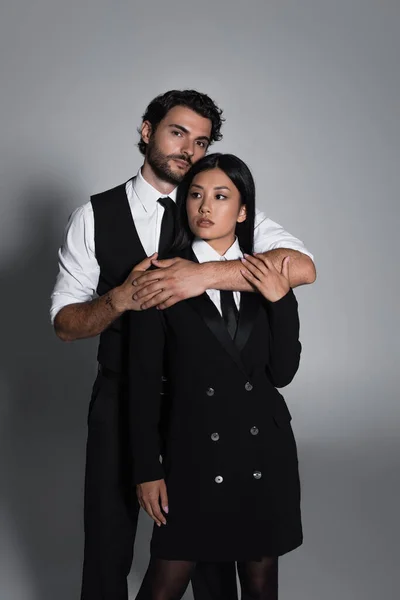 Elegante hombre abrazando a mujer asiática en negro blazer vestido sobre fondo gris - foto de stock