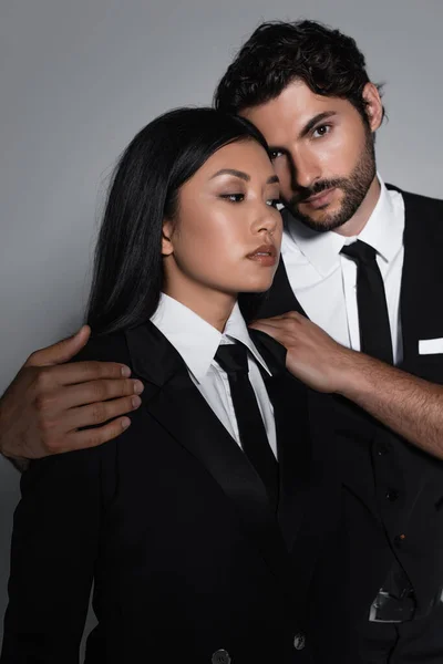 Элегантный брюнетка мужчина, смотрящий в камеру, обнимая чувственную азиатскую женщину, изолированную от серого — стоковое фото