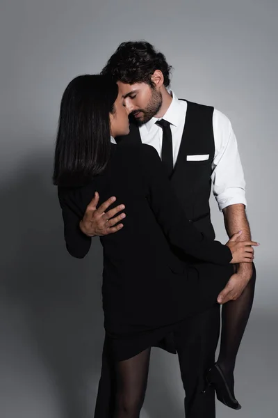 Élégant homme embrassant passionné asiatique femme en noir blazer robe sur fond gris — Photo de stock