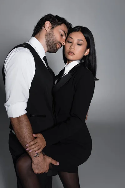 Elegante morena hombre seducir a mujer asiática en negro blazer vestido sobre fondo gris - foto de stock