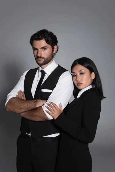 Elegante hombre de pie con brazos cruzados cerca de joven mujer asiática sobre fondo gris - foto de stock