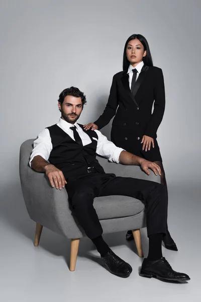 Elegante hombre sentado en sillón cerca de mujer asiática en negro blazer vestido sobre fondo gris - foto de stock