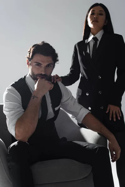 Brunette stylish man sitting in armchair near elegant asian woman on grey background — Stock Photo