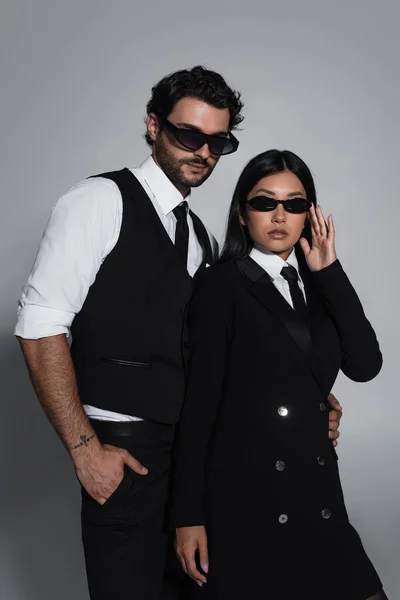 Elegante pareja interracial en ropa formal y gafas de sol negras mirando a la cámara sobre fondo gris - foto de stock