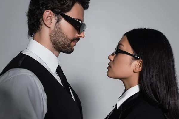 Vista laterale di coppia interrazziale in occhiali da sole neri guardando l'un l'altro su sfondo grigio — Foto stock