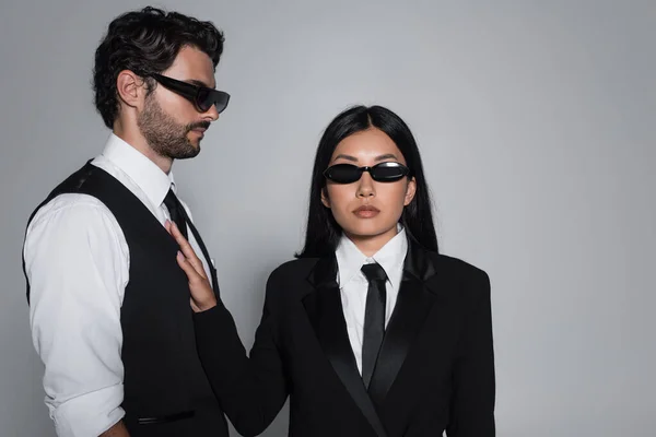 Élégante asiatique femme en noir formelle usure et lunettes de soleil regarder caméra près élégant homme isolé sur gris — Photo de stock