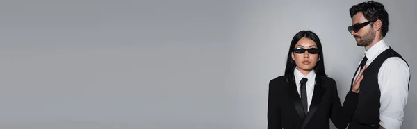 Donna asiatica in occhiali da sole neri e abbigliamento formale guardando la fotocamera vicino all'uomo alla moda isolato sul grigio, banner — Foto stock