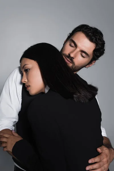 Uomo con gli occhi chiusi abbracciare sensuale asiatico donna isolato su grigio — Stock Photo