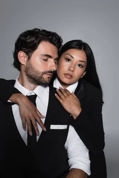 Hübsche asiatische Frau umarmt eleganten Mann und schaut auf Kamera isoliert auf grau — Stockfoto