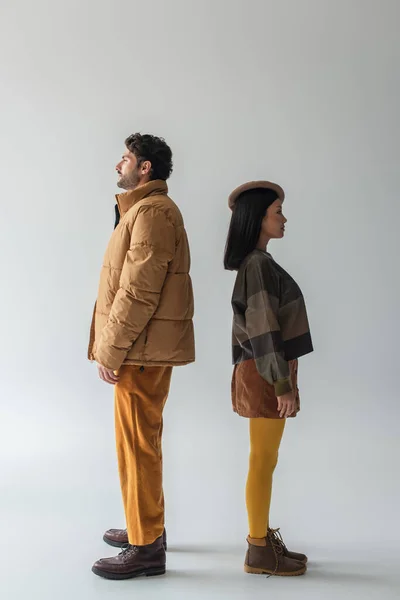 Full length view of interracial couple in beige and yellow clothes standing back to back on grey — Stock Photo