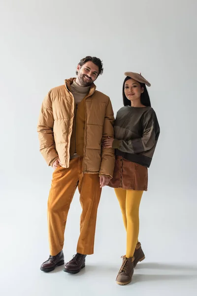 Vue pleine longueur du couple interracial en vêtements à la mode souriant à la caméra sur gris — Photo de stock