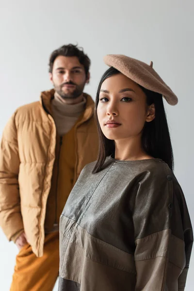 Asiatique femme en béret et en cuir pull regarder caméra près flou homme isolé sur gris — Photo de stock