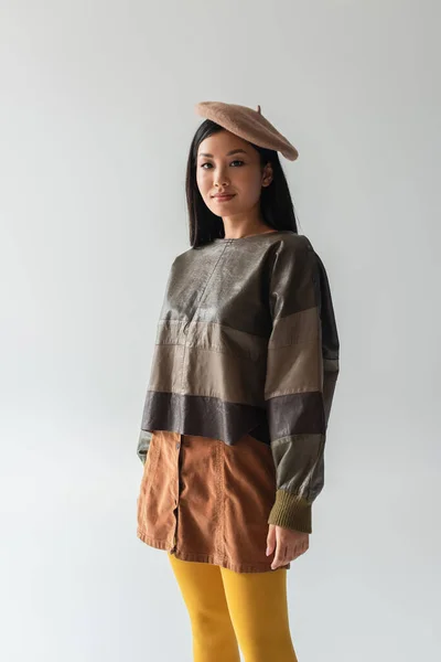 Молодая азиатка в кожаном свитере, бежевой юбке и берете смотрит на камеру, изолированную на сером — стоковое фото