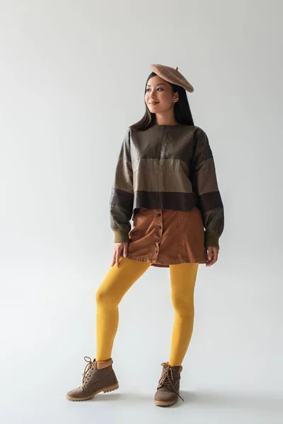 Piena vista lunghezza di donna asiatica sorridente in abiti alla moda e collant gialli guardando lontano sul grigio — Foto stock